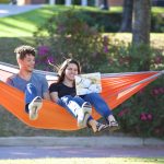 One man and one woman sitting on an orange hammock, smiling and looking at a laptop.