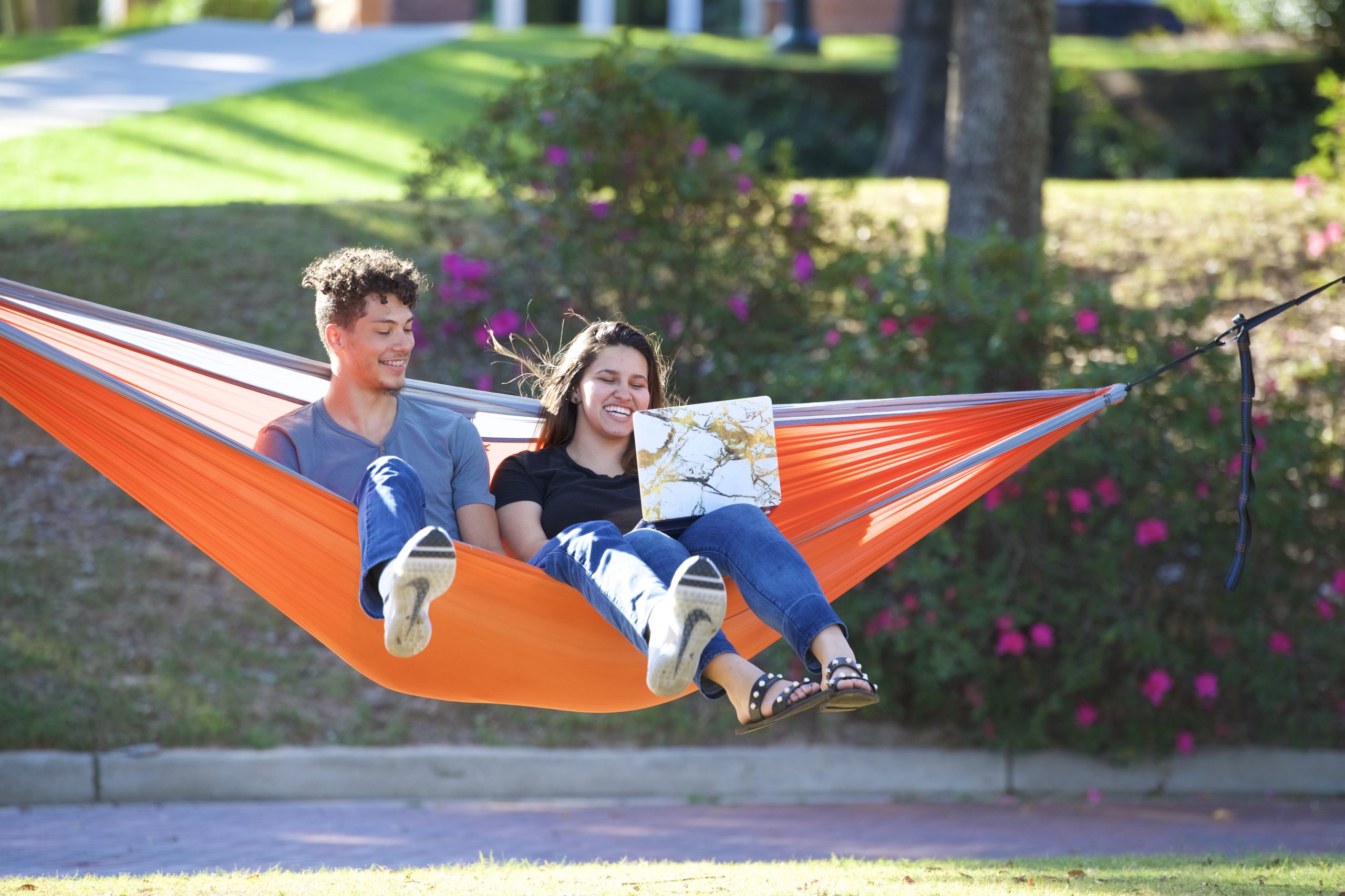 One man and one woman sitting on an orange hammock, smiling and looking at a laptop.